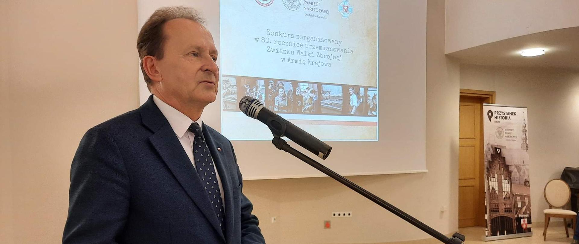 Wiceminister Włodzimierz Bernacki przemawia podczas gali konkursu. Stoi przy mikrofonie. Za nim wyświetlana jest prezentacja.