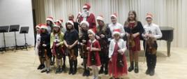Na zdjęciu grupa uczniów, którzy trzymają skrzypce i mają założone czerwone czapeczki mikołajkowe. Za nimi stoi w czerwonym stroju Święty Mikołaj.