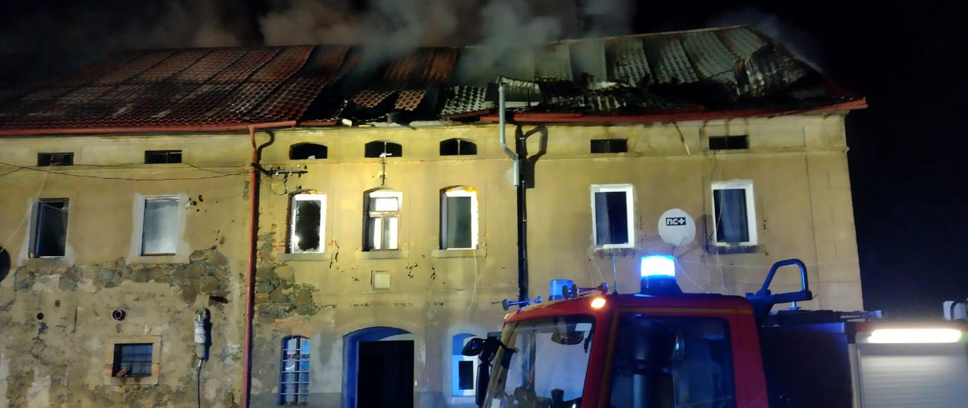 Pora nocna. Widok na budynek mieszkalny. Dach pokryty blachodachówką w kolorze ceglastym. Część blachy widoczne zapadnięta i okopcona. Z pomiędzy blachy wydobywa się biały dym. W centralnej części budynku znajduje się otwór wejściowy, nie widać drzwi. Przez otwór wejściowy przechodzą dwa węże strażackie. Przed budynkiem stoi samochód ratowniczo-gaśniczy, który na drzwiach w kolorze czerwonym ma numer operacyjny 601D51 w kolorze białym. Samochód ma włączone światła ostrzegawcze w kolorze niebieskim