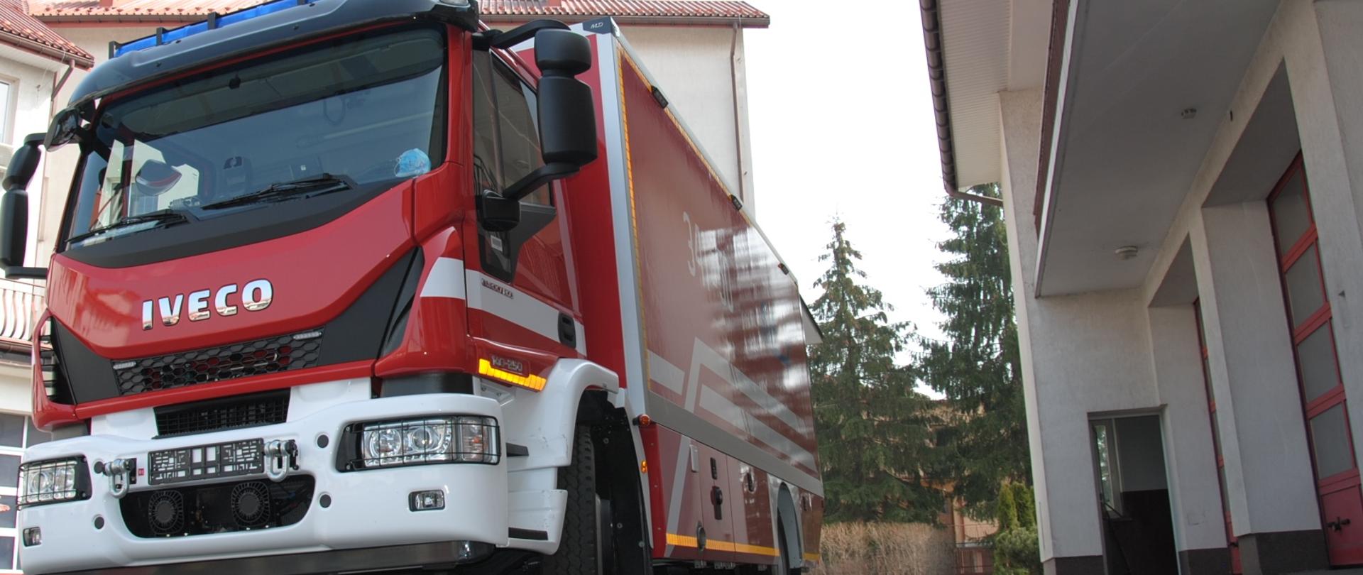 Zdjęcie przedstawia przód samochodu pożarniczego marki IVECO koloru czerwonego z białym zderzakami i białymi pasami na drzwiach i tylnej zabudowie. Na dachu posiada zamontowaną belkę z niebieskimi światłami błyskowymi do poruszania się w ruchu uprzywilejowanym. Pojazd stoi na placu wewnętrznym Komendy Miejskiej Państwowej Straży Pożarnej. W tle widoczne są budynki Komendy.