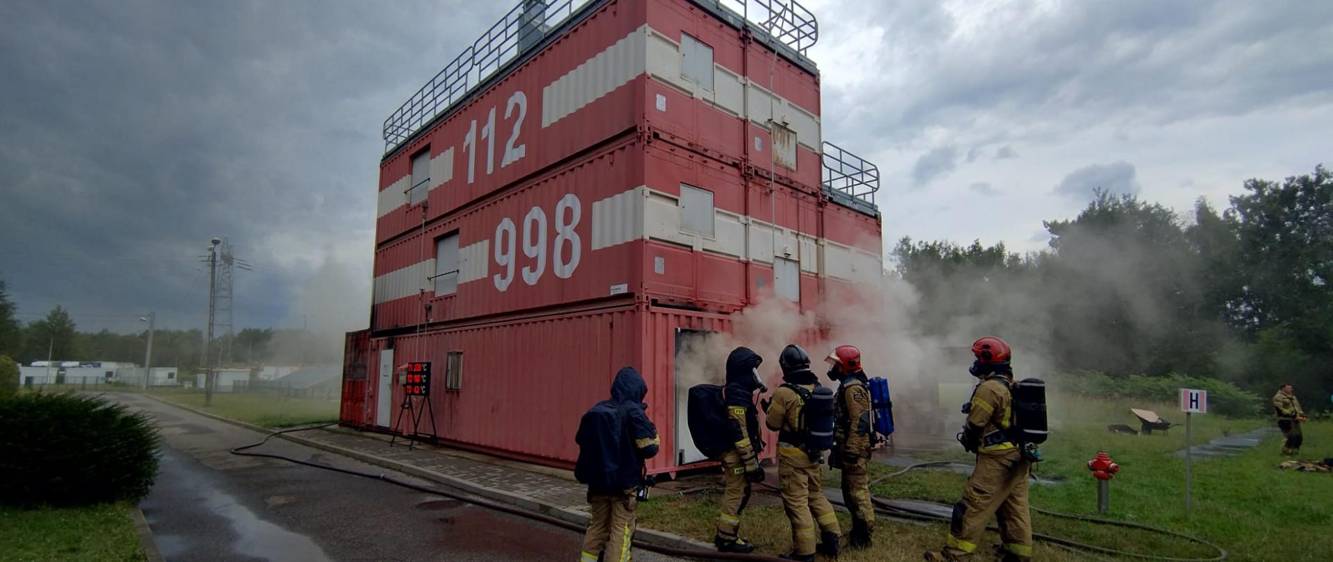 Poligon ćwiczebny. Kilku strażaków w umundurowani bojowym stoi przed kontenerem rozgorzeniowym