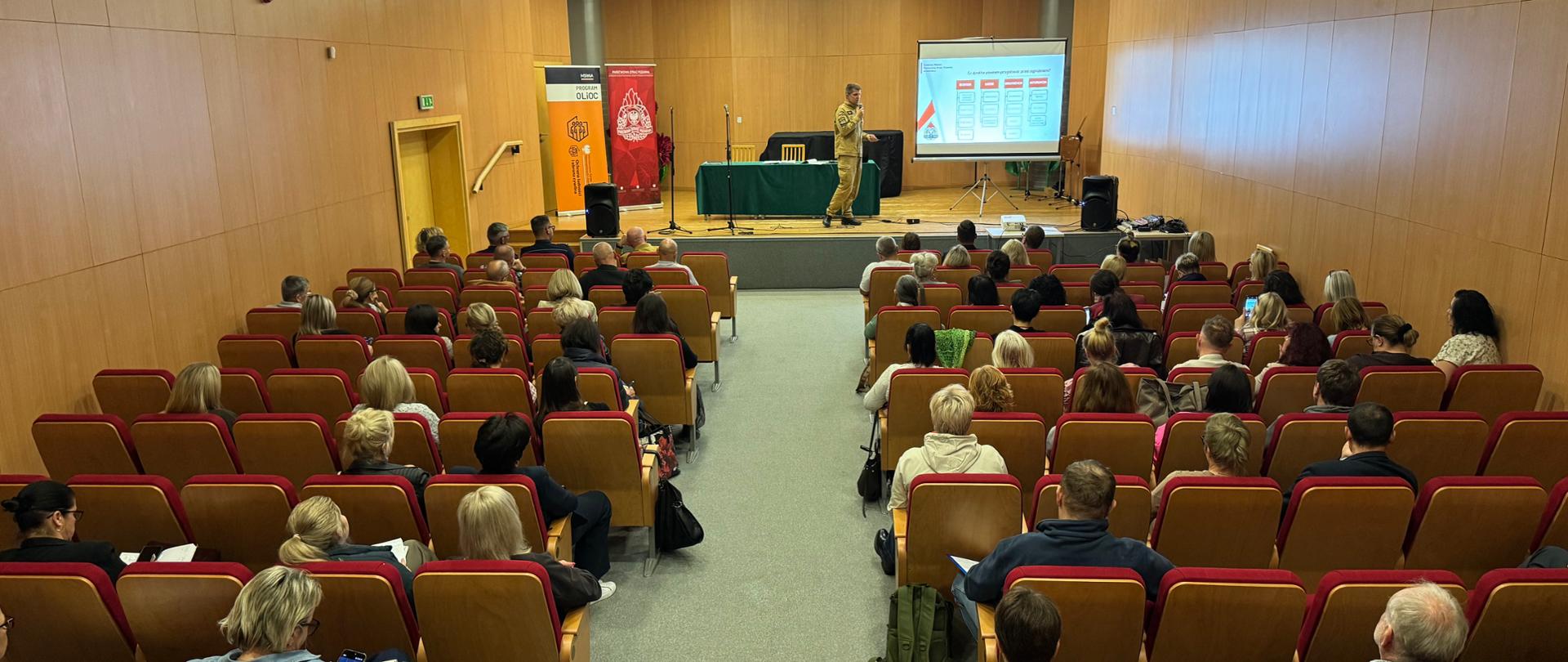Sala konferencyjna z drewnianymi ścianami i rzędami czerwono-brązowych foteli, w których siedzą uczestnicy spotkania. Na podwyższeniu znajduje się prelegent w piaskowym mundurze PSP, obok stołu przykrytego zielonym obrusem i ekranu z prezentacją multimedialną. Po bokach sceny stoją roll-upy promocyjne. Uczestnicy zwróceni są w stronę prowadzącego, a szeroki korytarz pośrodku sali prowadzi do sceny.