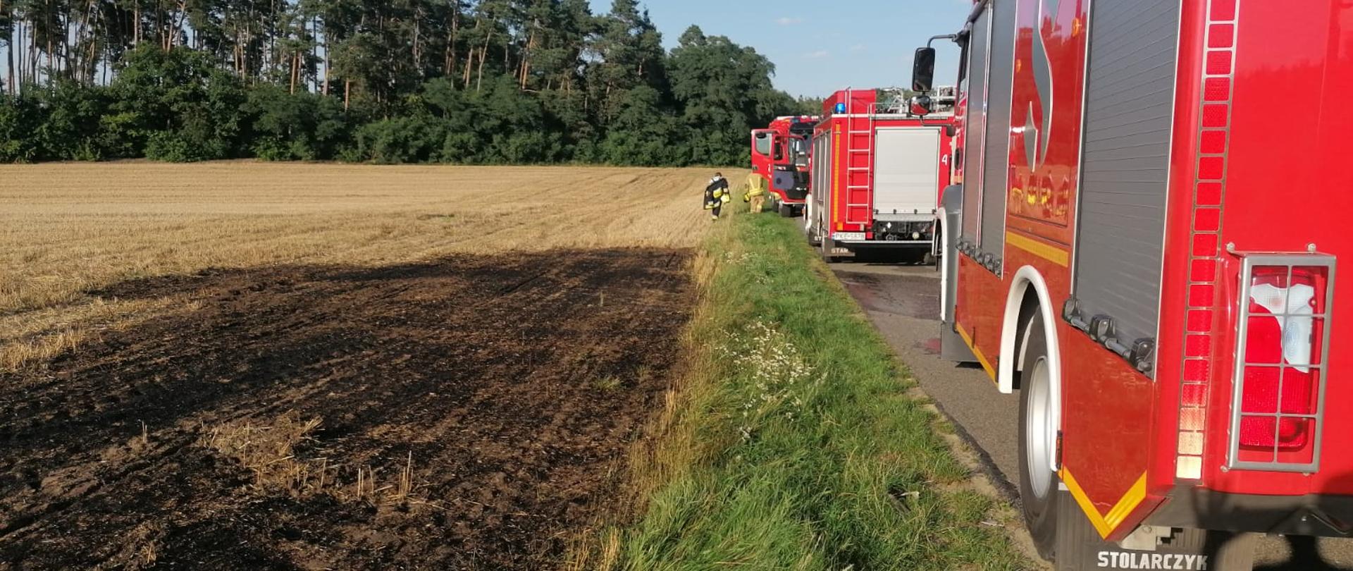 Widoczne miejsce zdarzenia pojazdy pożarnicze oraz wypalone ściernisko.