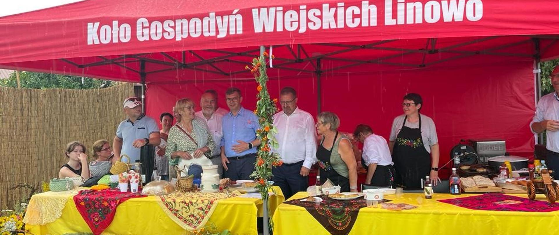 Pod czerwonym namiotem z napisem Koło Gospodyń Wiejskich Linowo udekorowane snopami słomy, żółtymi obrusami i ludowymi chustami stoły, w dalszym planie gospodynie z gośćmi odwiedzającymi stoisko