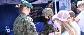 Żołnierka w mundurze pomaga dziewczynce przymierzyć kamizelkę taktyczną i hełm na stoisku promocyjnym Wojska Polskiego. W tle widoczny napis „Dobrowolna Zasadnicza Służba Wojskowa”.