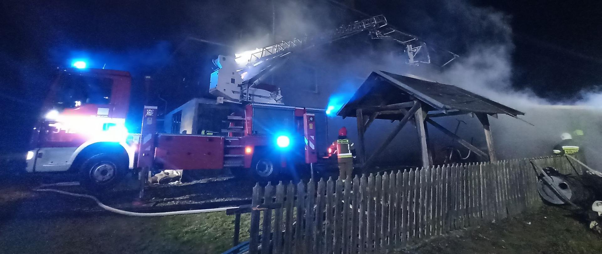 Zdjęcie przedstawia działania ratowniczo-gaśnicze prowadzone przez strażaków z wykorzystaniem pożarniczego podnośnika hydraulicznego. Nad spalonym budynkiem gospodarczym unosi się dym i para.