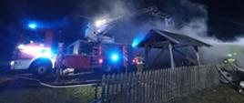 Zdjęcie przedstawia działania ratowniczo-gaśnicze prowadzone przez strażaków z wykorzystaniem pożarniczego podnośnika hydraulicznego. Nad spalonym budynkiem gospodarczym unosi się dym i para.