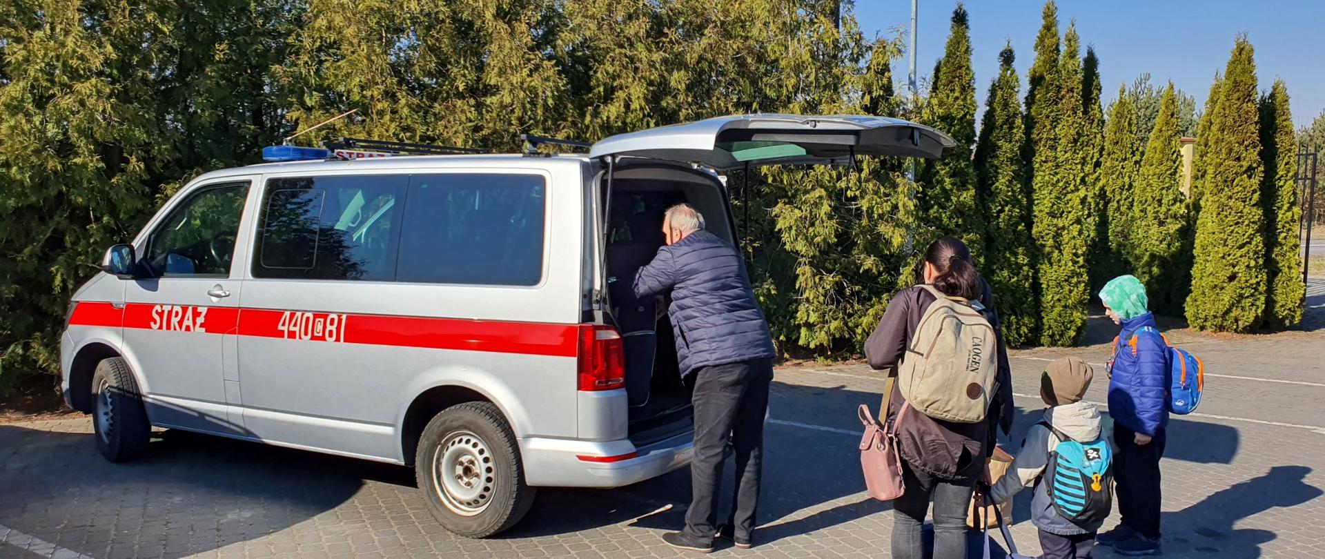 Zdjęcie przedstawia kobietę z dwójką dzieci i mężczyznę widzianych od tyłu. Mężczyzna układa torby w otwartym bagażniku pojazdu stojącego na utwardzonym placu. W tle zielone krzewy.