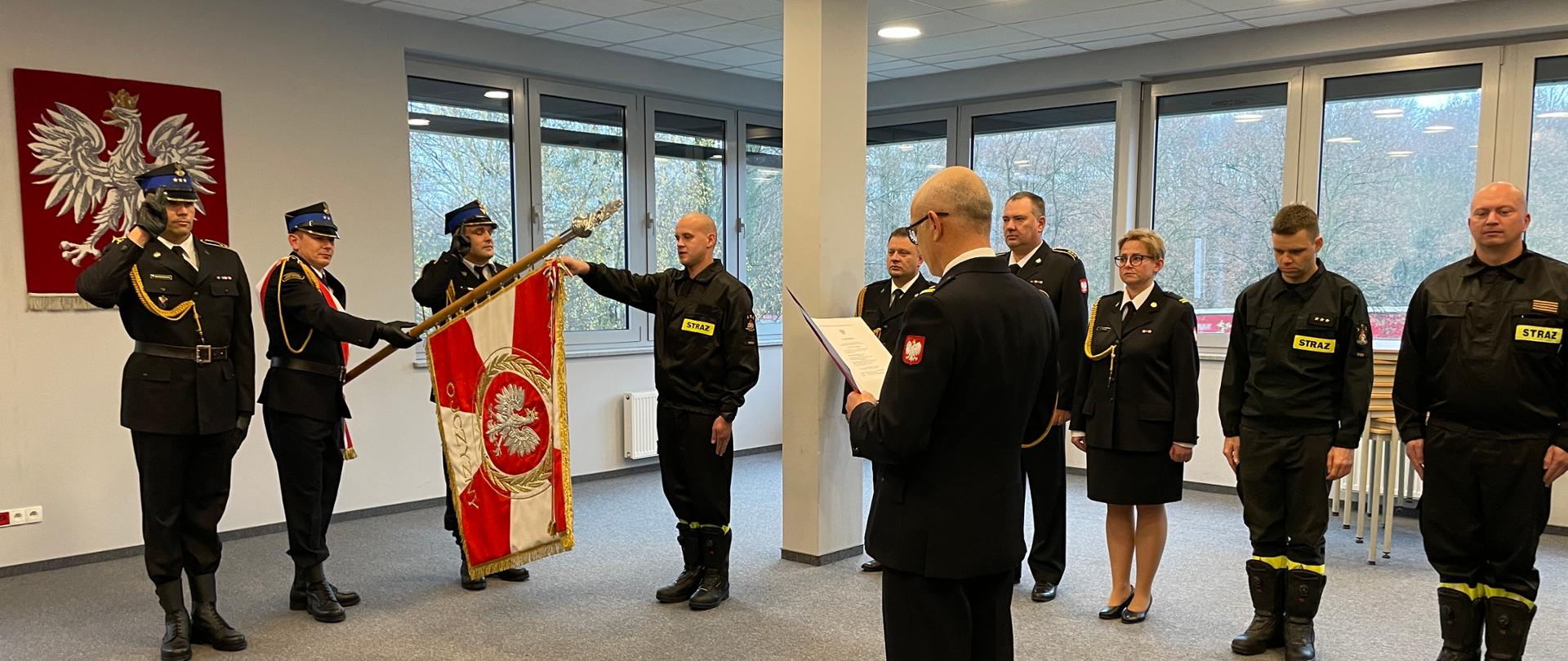 Po lewej stronie stoi poczet sztandarowy z opuszczonym sztandarem podczas składania ślubowania. W centralenj części zdjęcia stoi Komendant Powiatowy PSP w Śremie. Po prawej stronie stoją funkcjonariusze obecni podczas ślubowania 