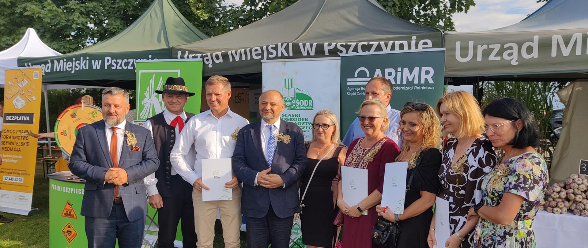 Dożynki w Łące, 30 sierpnia, zdjęcie uczestników - grupa 10 osób, stojących w rzędzie do fotografii, w tle namioty firmowe z logotypami