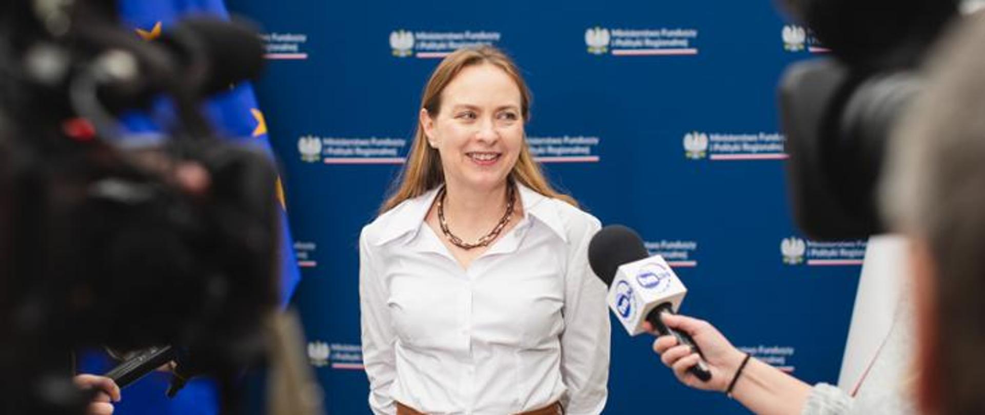 Minister Katarzyna Pełczyńska-Nałęcz podczas konferencji prasowej, widoczne mikrofony i kamery.