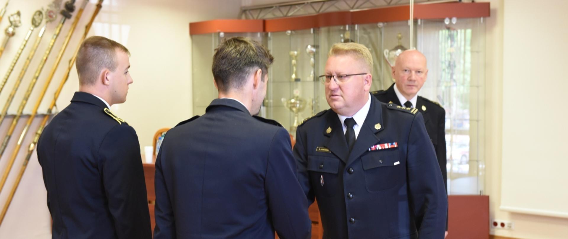 Na zdjęciu widać 4 strażaków w sali, komendant wojewódzki PSP gratuluje 1 ze strażaków