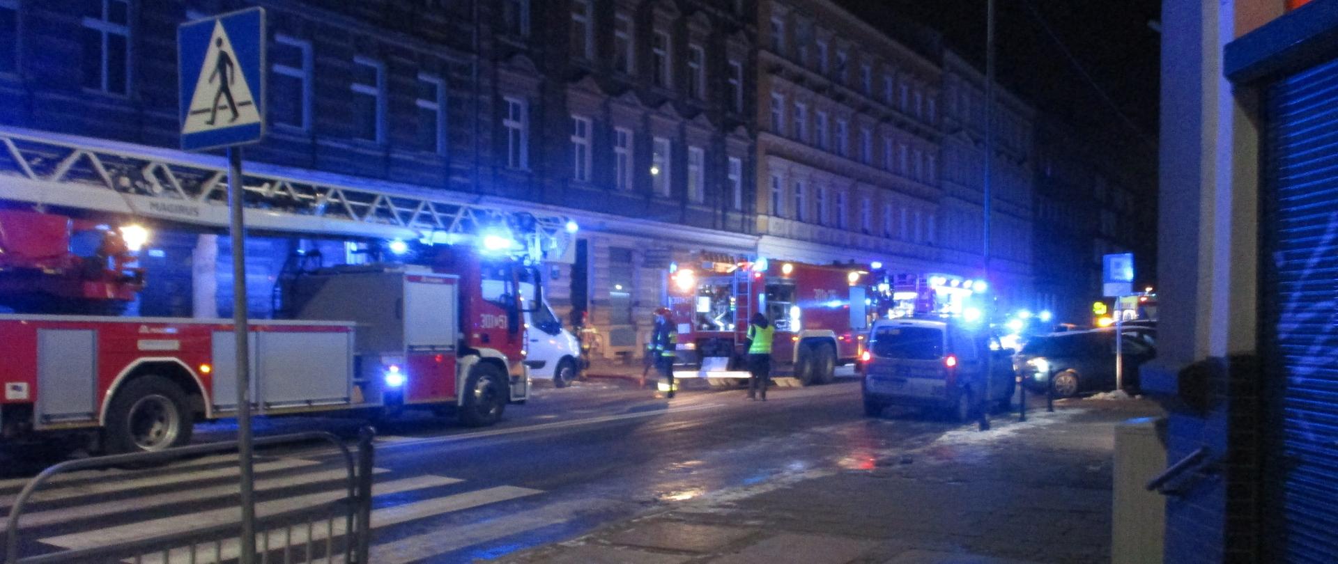 Zdjęcie obejmuje chodnik, ulicę, przejście dla pieszych oraz samochody straży pożarnej i straży miejskiej w tle widać kamienice przy ulicy Śląskiej. Linia gaśnicza z środkowego pojazdu PSP prowadzona w przejście bramowe. Widać pracujących dwóch strażaków oraz strażnika miejskiego.