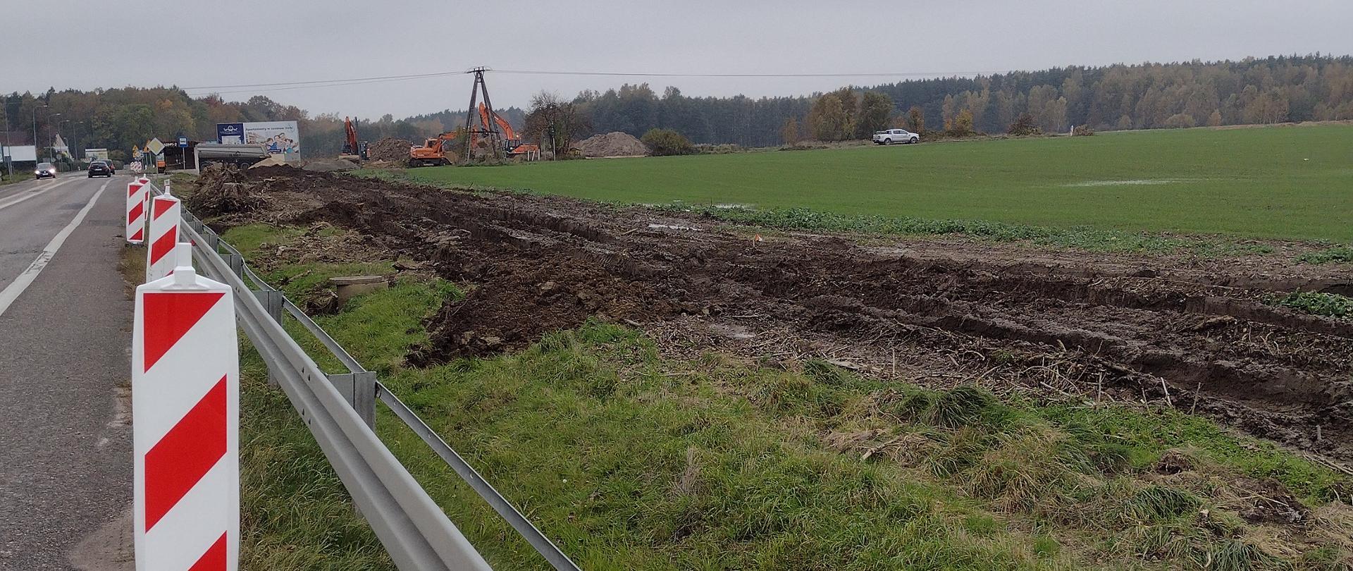 Zdjęcie przedstawia rozbudowywaną drogę krajową nr 21 między Słupskiem a Ustką i pobocze z widocznymi pracami ziemnymi oraz pracującym w oddali sprzętem budowlanym.