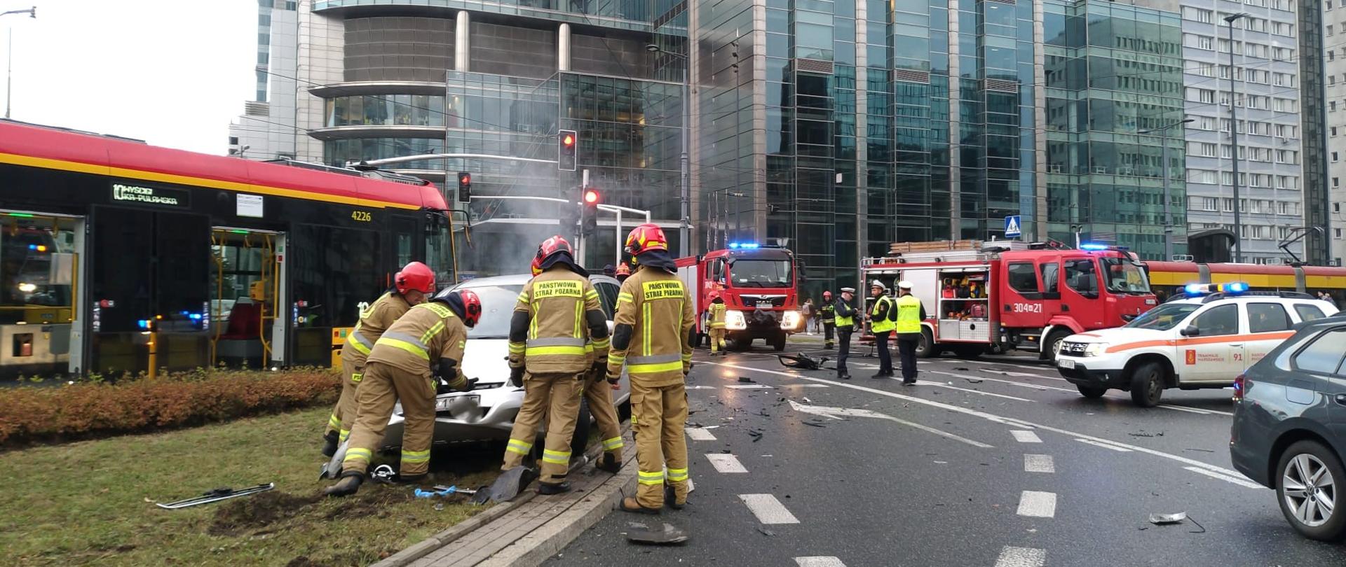 Na fotografii widać dwa czerwone samochody strażackie , Nadzór Ruchu Tramwaje Warszawskie oraz tramwaj koloru żółtego , który brał udział w zdarzeniu. Na zdjęciu widać też strażaków w ubraniach ochronnych, którzy prowadzą działania.