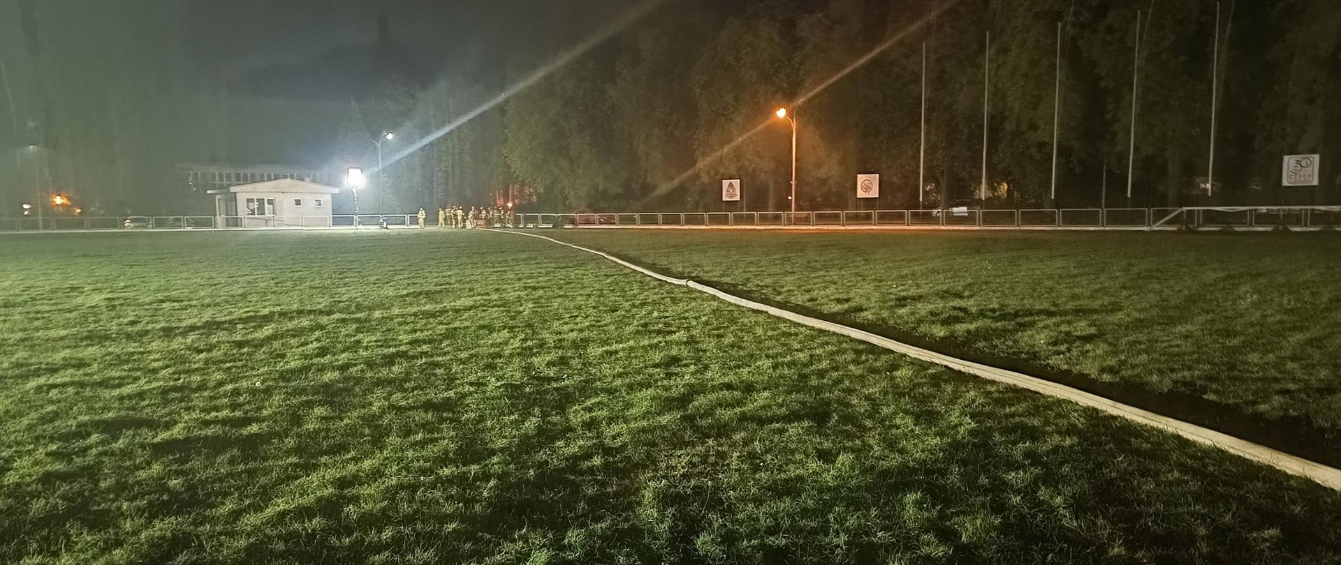 Pora nocna, murawa stadionu przez którą przebiega rozciągnięta linia wodna, w głębi widać stojącą grupę strażaków
