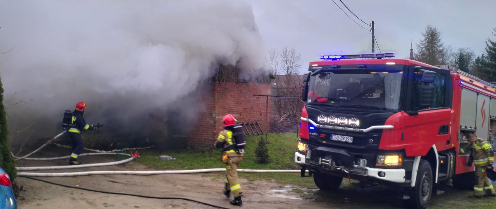 Fotografa przedstawia widok budynku gospodarczego, z którego wydobywają się duże ilości dymu, przy budynku strażacy podają prądy wody, obok widoczny pojazd pożarniczy i inni ratownicy wyciągający z niego sprzęt.