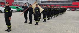 Na zdjęciu widzimy strażaków ustawionych w szyku w czarnych czapkach z daszkiem, czarnych mundurach, pasach głównych z toporkami oraz w czarnych butach typu bukaty podkute. Pododdział ćwiczy elementy uroczystości pożarniczych. Na zdjęciu widzimy również przed pododdziałem osoby szkolące również w mundurach strażackich.