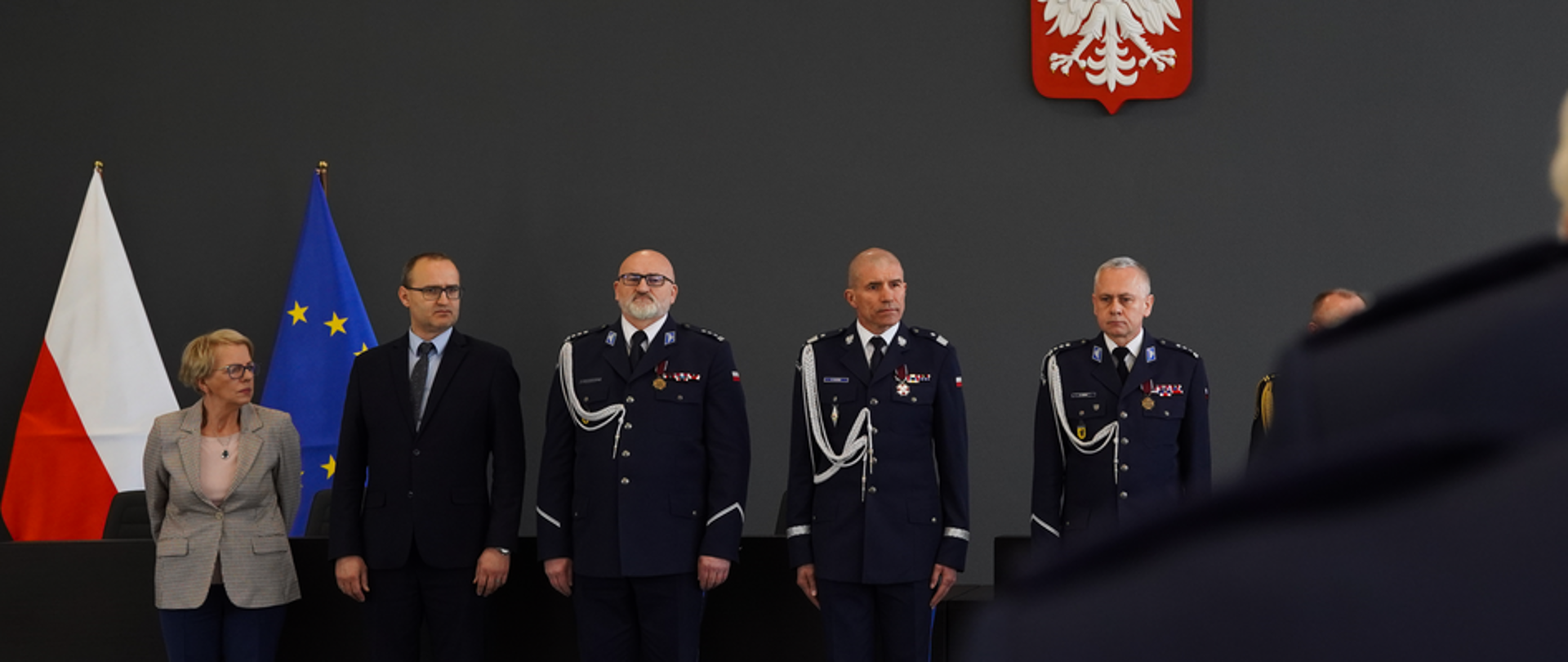 grupa mężczyzn w mundurach Policji , mężczyzna w garniturze i kobieta stoją przy ścianie na której wisi godło Polski. 