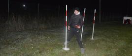 Biwak klas mundurowych. Zdjęcie przedstawia teren biwaku, na którym odbywa się nocny sprawnościowy tor przeszkód. Jeden z uczestników "zalicza" slalom między tyczkami. W lewej ręce trzyma czerwoną pałkę świetlną. 