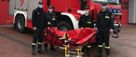 Zdjęcie przedstawia czterech strażaków stojących przed samochodem strażackim trzymających w ręku nosze płachtowe do transportu medycznego koloru czerwonego. Przed nimi stoi specjalistyczne krzesełko do transportu kardiologicznego. Strażacy ubrani są w czarne ubrania koszarowe, na twarzy maja maseczki a na głowie czapkę z daszkiem. W tle widnieje budynek Komendy Powiatowe PSP z dużym napisem Państwowa Straż Pożarna barwy czerwonej, widać bramy garażowe. 