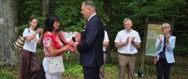 Zdjęcie przedstawia uroczysty moment wręczania kwiatów w leśnej scenerii