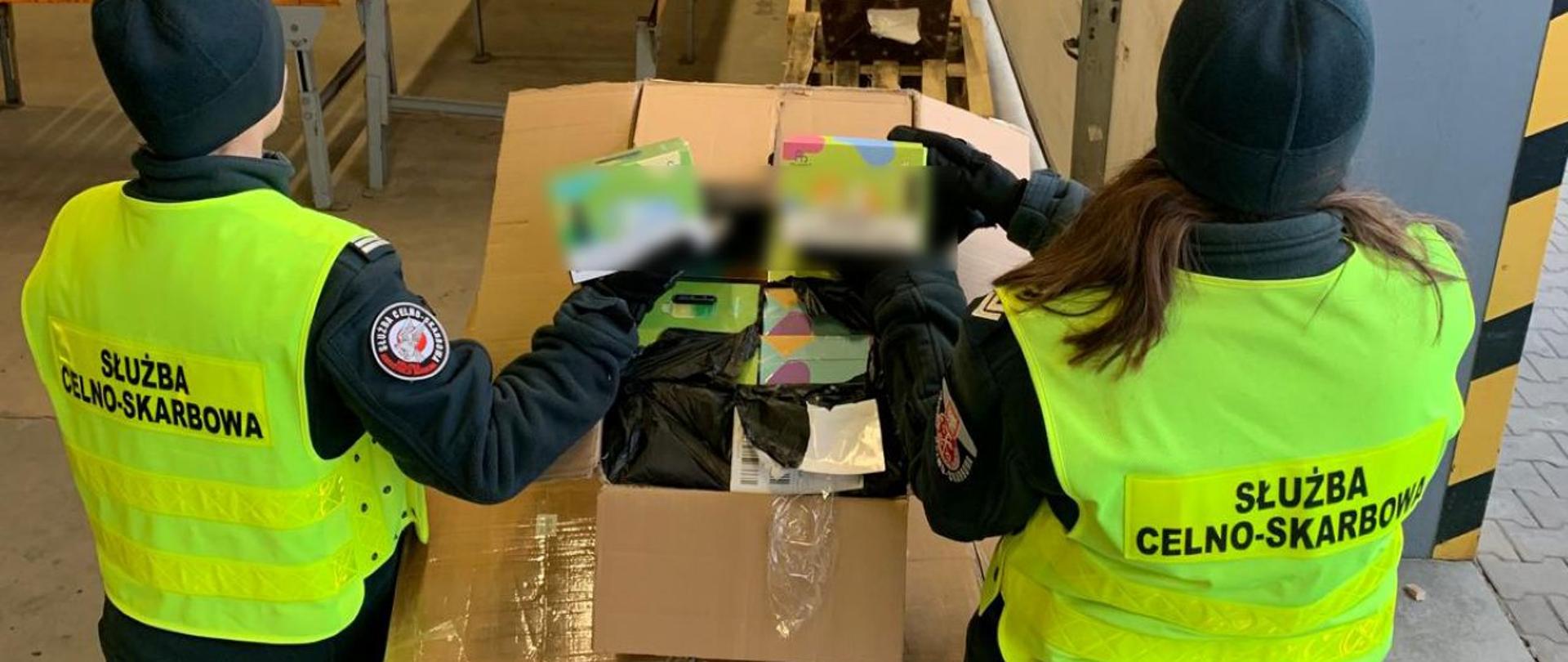 Dwóch funkcjonariuszy Służby Celno‑Skarbowej przegląda zawartość dużego kartonu w magazynie.