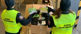 Dwóch funkcjonariuszy Służby Celno‑Skarbowej przegląda zawartość dużego kartonu w magazynie.
