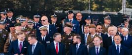Grupa przedstawicieli władz oraz wysokich rangą oficerów różnych służb mundurowych zgromadzona na trybunie podczas uroczystości państwowej. Uczestnicy stoją w kilku rzędach, część w mundurach galowych, a część w garniturach.