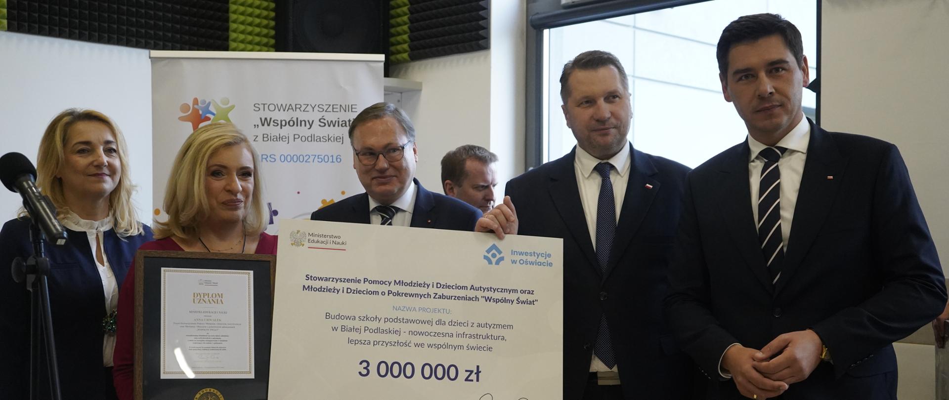 Minister Czarnek stoi w otoczeniu kilku osób, mężczyzna obok niego trzyma wielki symboliczny czek z napisem 3 000 000 zł.