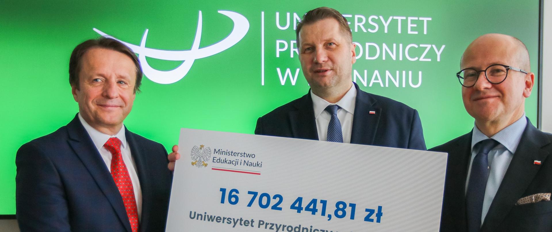 Minister Czarnek stoi pomiędzy dwoma mężczyznami w garniturach, jeden z nich trzyma wielki symboliczny czek z napisem 16 702 441,81 zł, za nimi zielona ściana z napisem Uniwersytet Przyrodniczy w Poznaniu.