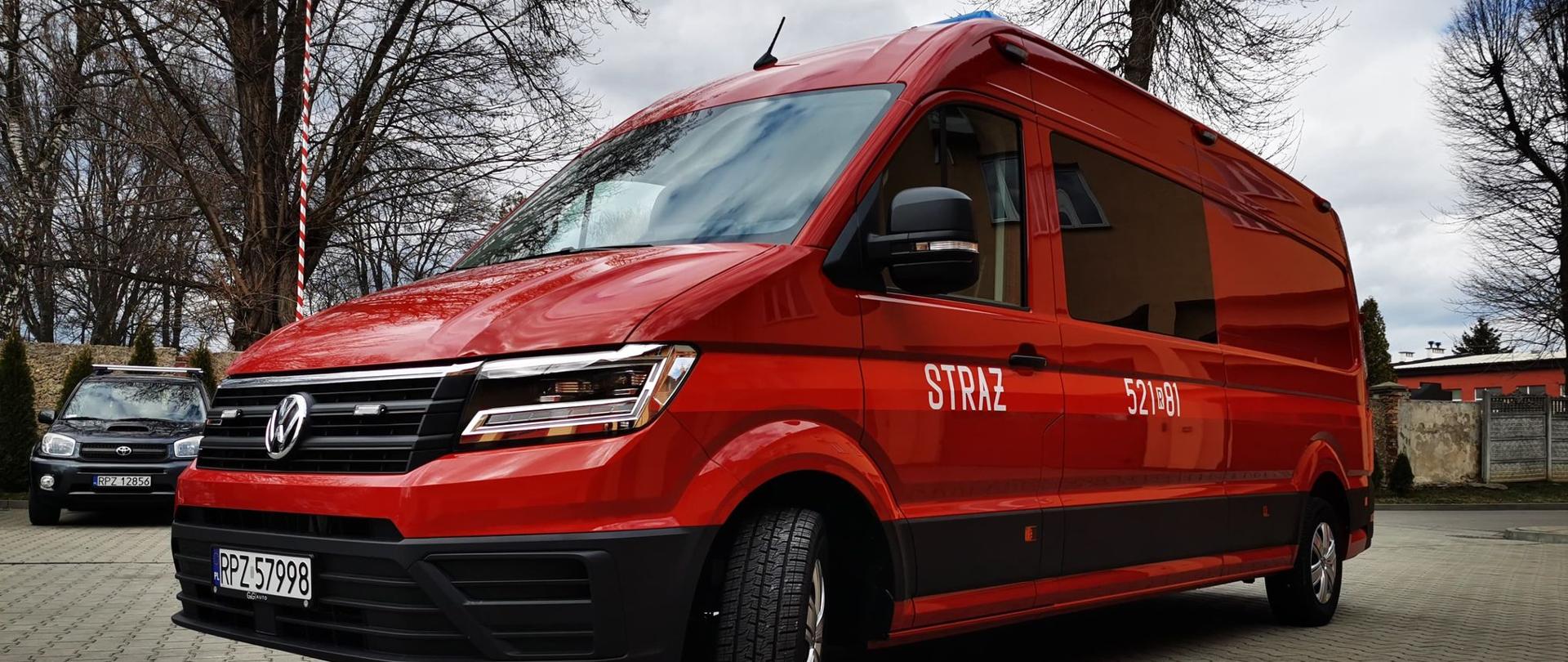 W centralnej części zdjęcia przedstawiony jest samochód - bus- do przewozu i działań zespołu dronowego w kolorze czerwonym marki Volkswagen zwrócony maską i jednym z boków do zdjęcia. Na karoserii drzwi kierowcy biały napis "Straż". Pojazd został sfotografowany w pochmurny dzień na parkingu. 