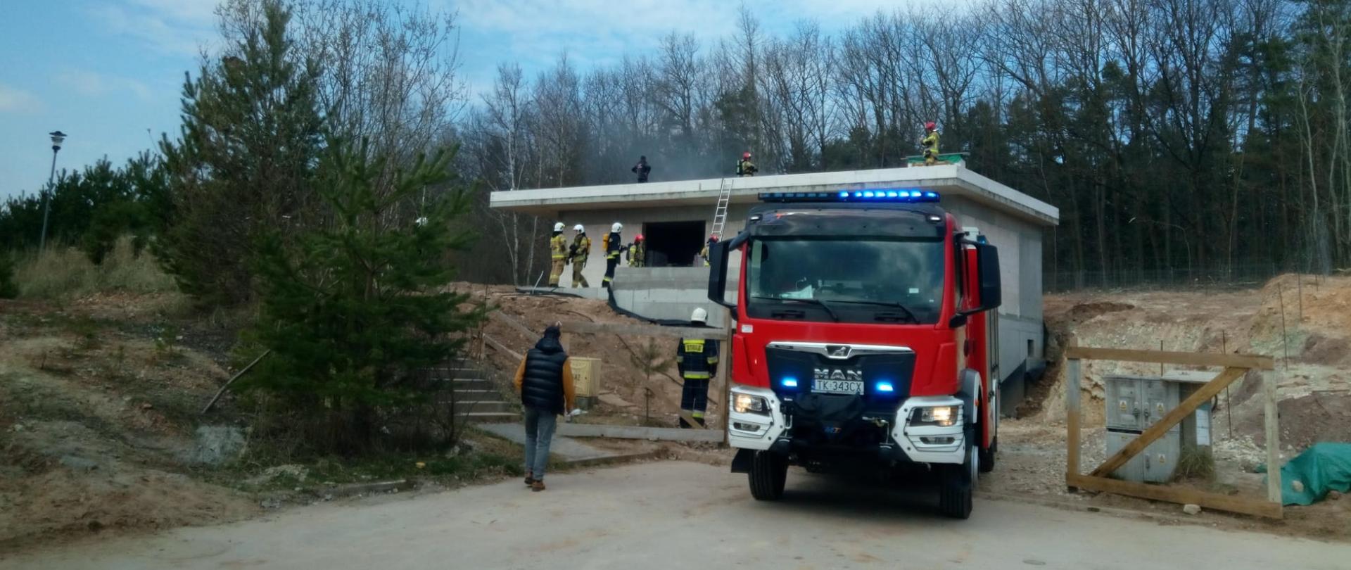 Zdjęcie przedstawia samochód pożarniczy stojący przy wjeździe na budowę gdzie doszło do pożaru dachu. Za pojazdem, po lewej strony widać pracujących strażaków, a w głębi nowo budowany dom.