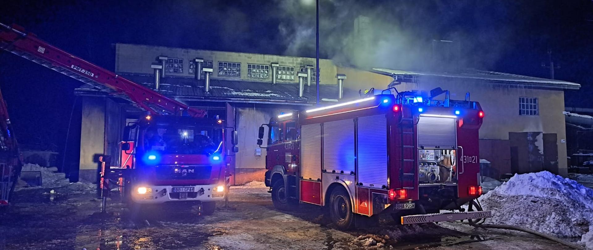 W nocy na zaśnieżonym podwórku przed budynkiem stoją dwa wozy strażackie z włączonymi sygnałami świetlnymi.