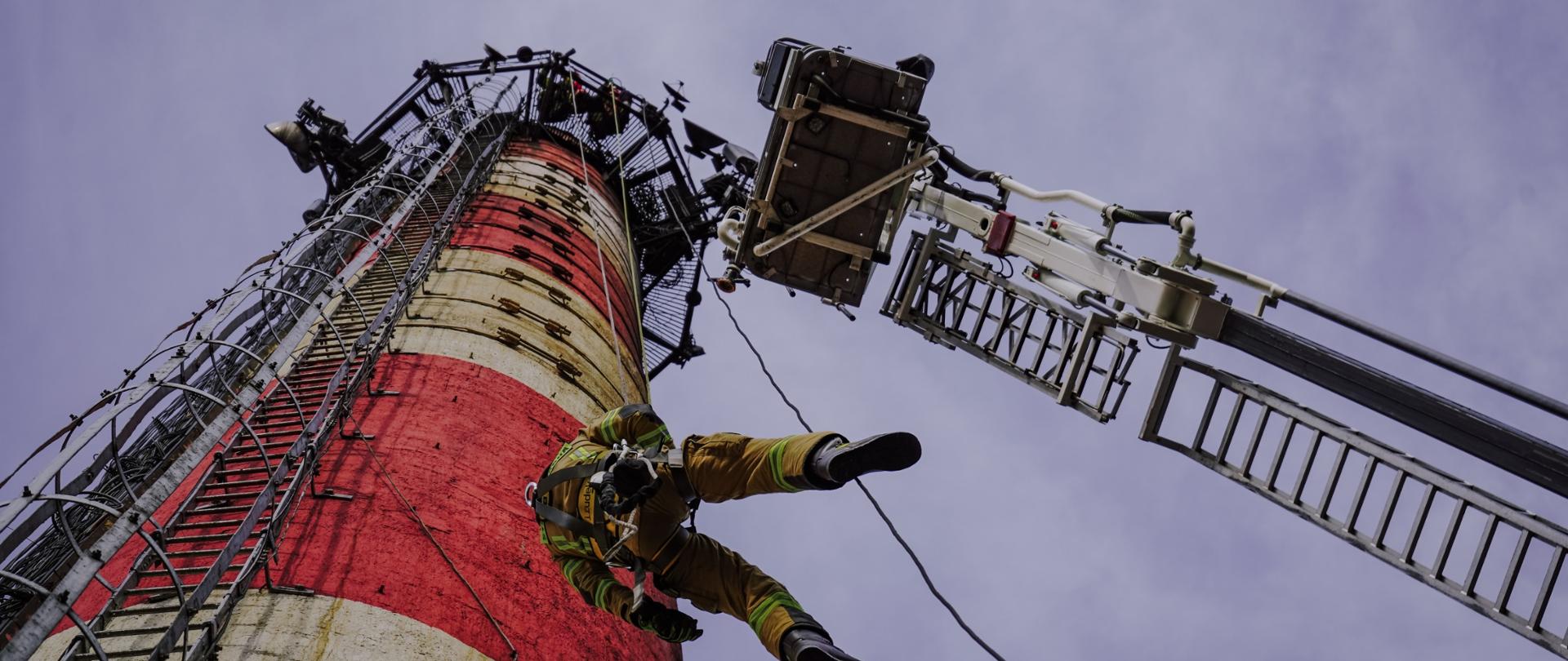 Strażacy ćwiczą zjazd z komina. Obok rozstawiony podnośnik hydrauliczny.