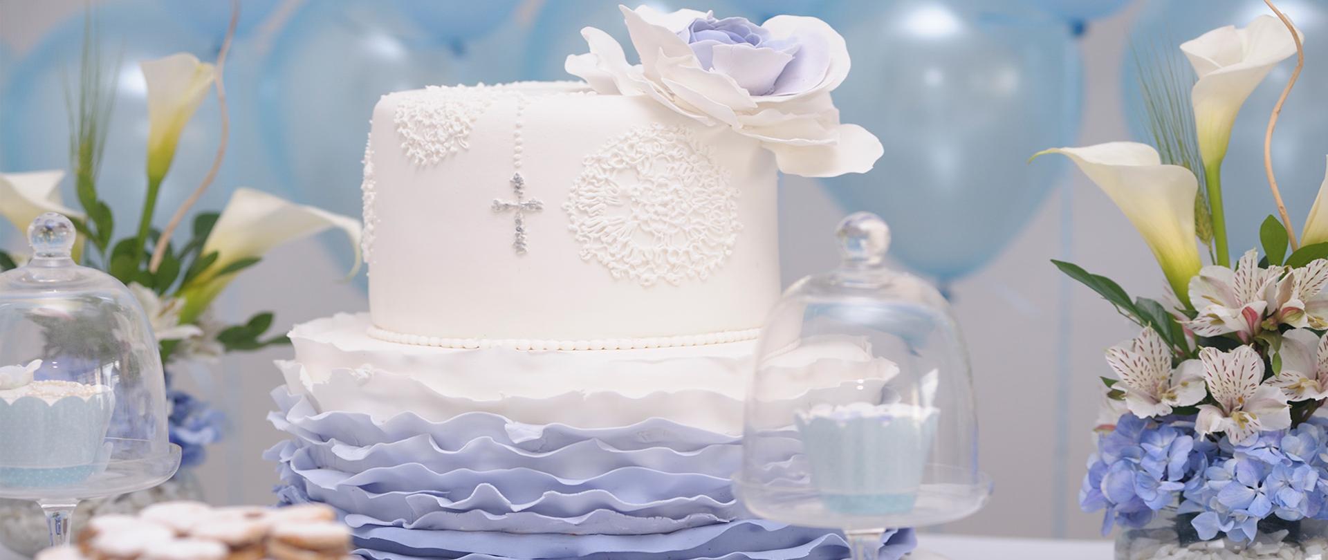 Na zdjęciu stół zastawiony kwiatami i daniami komunijnymi takimi jak tort z kremem, babeczki i ciasteczka. 