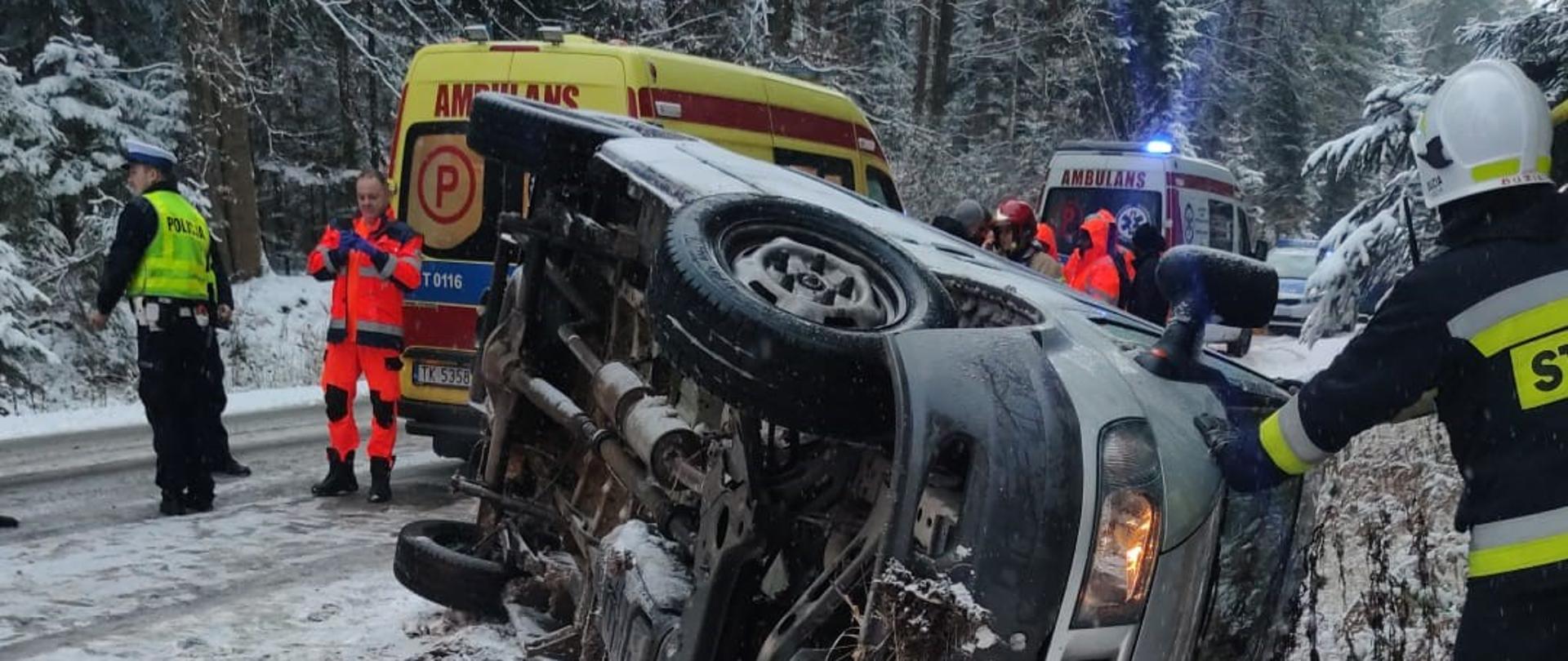 Zdjęcie przedstawia samochód osobowy, który leży na boku w przydrożnym rowie. Na jezdni za pojazdem widać dwa ambulanse Państwowego Ratownictwa Medycznego.