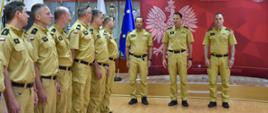 Grupa mężczyzn w mundurach stoi w rzędzie. W tle widoczna jest czerwona ściana z logo Państwowej Straży Pożarnej i napisem "PAŃSTWOWA STRAŻ POŻARNA KOMENDA WOJEWÓDZKA PSP W ŁODZI"