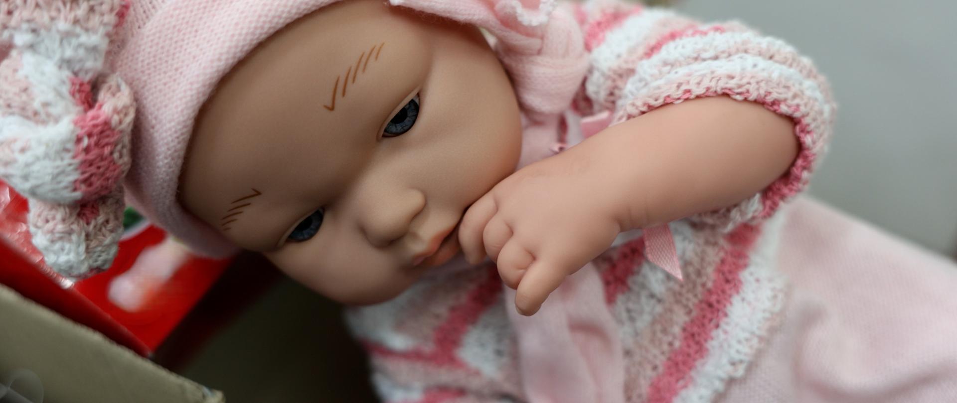Zbliżenie na lalkę stylizowaną na niemowlę, ubraną w różową czapkę i dzianinowy strój, leżącą w pudełku z ręką przy twarzy
