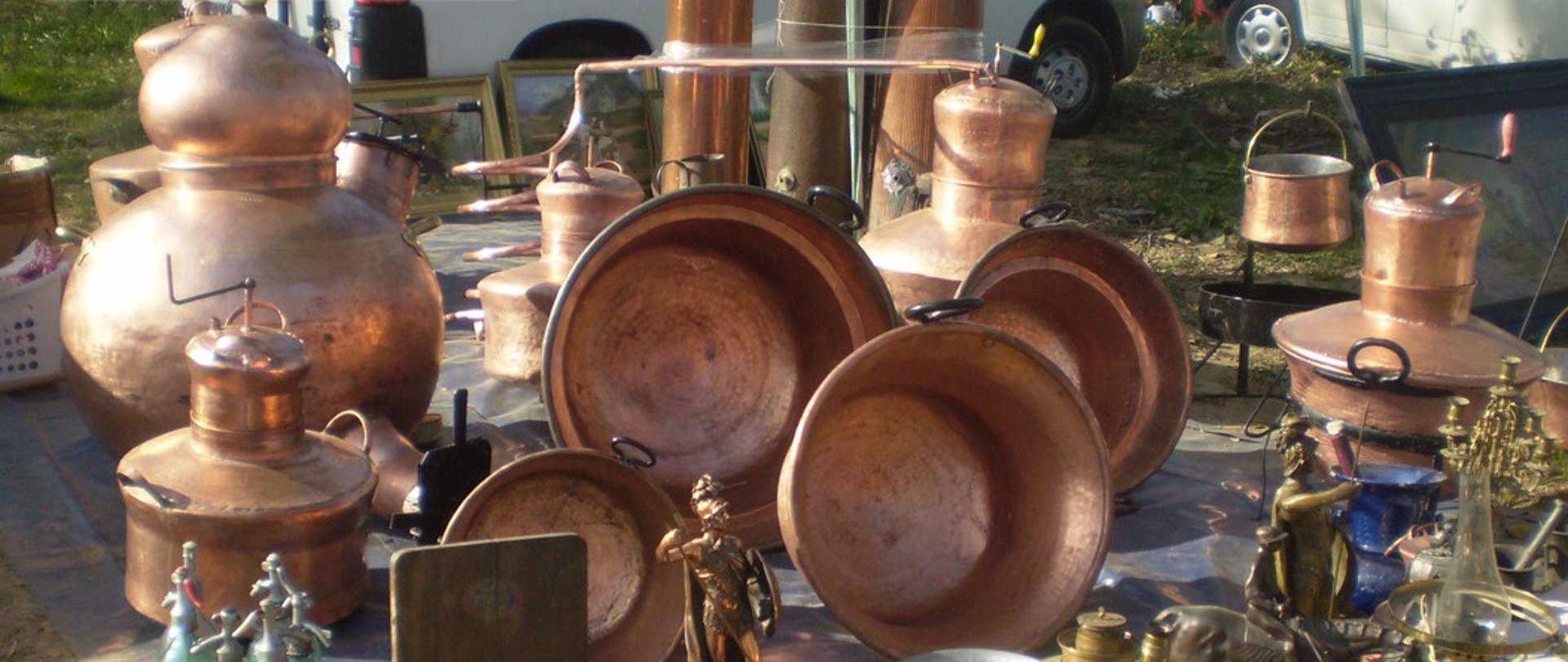 Zdjęcie przedstawia wyroby romskie - naczynia miedziane, kotły , butelki syfonów, figurki. Na pierwszym planie duża waga