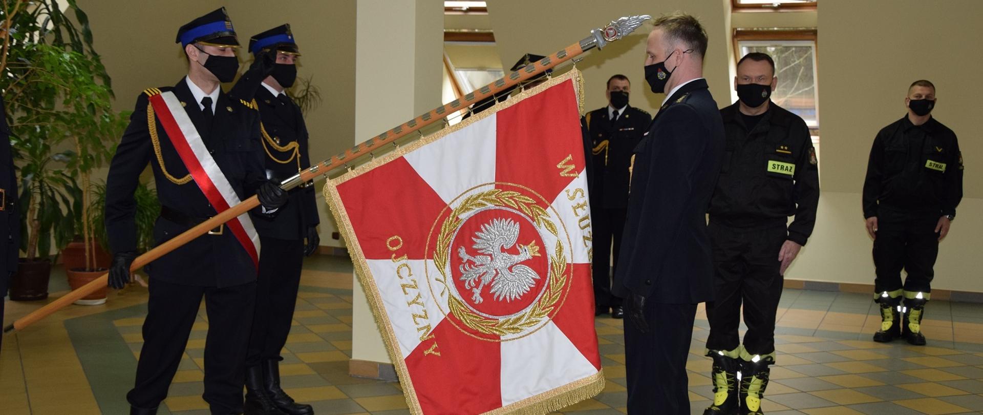 Na zdjęciu widać strażaka stojącego przed sztandarem, który trzyma inny strazak. W tle strażacy ustawieni na zbiórce.