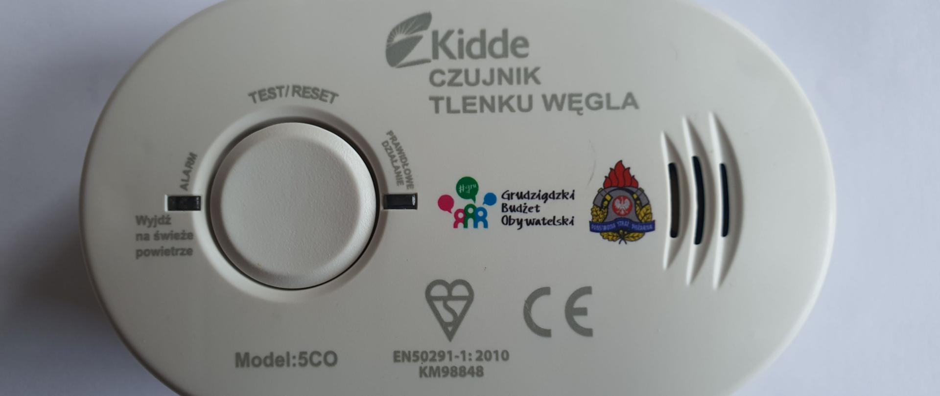 Zdjęcie przedstawia białą czujkę tlenku węgla w kształcie owalu, na której znajdują się napisy, diody informacyjne i przycisk funkcyjny.