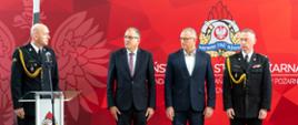 Na zdjęciu widoczna jest uroczysta zbiórka na sali gimnastycznej Komendzie Wojewódzkiej Państwowej Straży Pożarnej w Katowicach związaną z powitaniem powołanego zastępcy śląskiego komendanta wojewódzkiego. Na pierwszym planie, po lewej stronie, przy przezroczystej mównicy z logo Państwowej Straży Pożarnej, stoi mężczyzna w wyjściowym mundurze strażackim. Ma przypięte odznaczenia i sznur galowy w kolorze złotym przewieszony przez ramię. Przemawia do mikrofonu jest to zastępca śląskiego komendanta wojewódzkiego mł. bryg. Zbigniew Dyk. Obok niego stoją trzej mężczyźni – dwóch w garniturach (jeden w ciemnym garniturze i białej koszuli, drugi w garniturze i krawacie w drobne wzorki) Pan Grzegorz Mucha – Dyrektor Wydziału Bezpieczeństwa i Zarządzania Kryzysowego Śląskiego Urzędu Wojewódzkiego, Pan Marek Misiura – pełniący obowiązki Dyrektora Biura Bezpieczeństwa Urzędu Marszałkowskiego oraz strażak w mundurze wyjściowym – komendant wojewódzki st. bryg. Damian Legierski, również z odznaczeniami i złotym sznurem na ramieniu. Wszyscy patrzą w kierunku przemawiającego. Tło stanowi czerwony baner z dużym, stylizowanym orłem i herbem Państwowej Straży Pożarnej – przedstawiającym hełm strażacki, liście dębowe i płomienie, a w środku biały orzeł w czerwonym polu. W tle widoczne są także dane kontaktowe Komendy Wojewódzkiej Państwowej Straży Pożarnej w Katowicach.