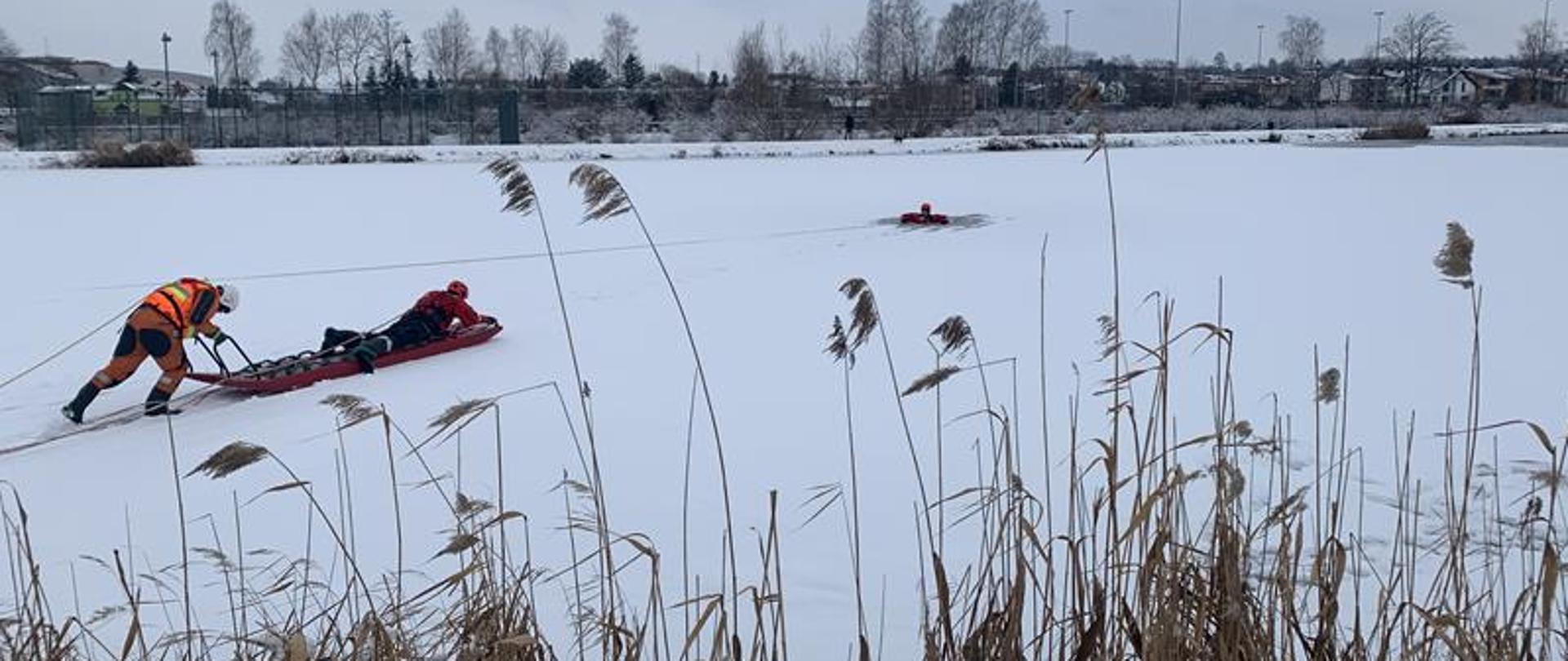 Strażacy ubrani w suche skafandry. Ćwiczą na zamarzniętym zalewie. Jeden znajduje się na saniach lodowych drugi natomiast pcha sanie na zamarzniętym zalewie.