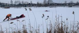Strażacy ubrani w suche skafandry. Ćwiczą na zamarzniętym zalewie. Jeden znajduje się na saniach lodowych drugi natomiast pcha sanie na zamarzniętym zalewie.