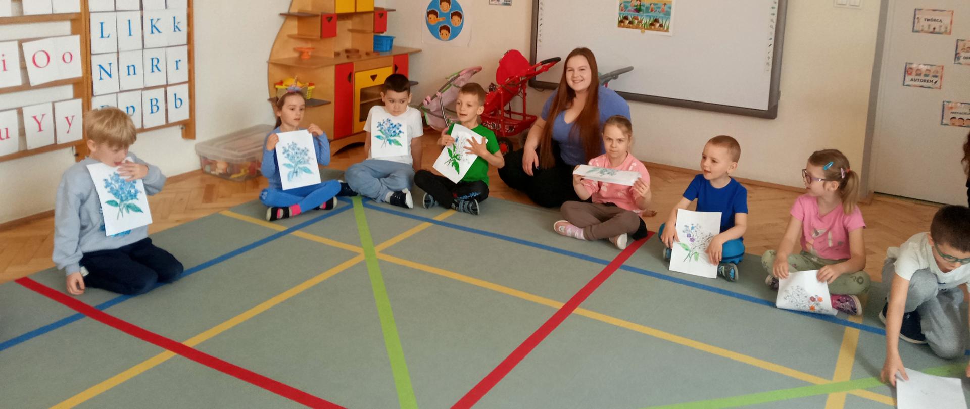 Dzieci pozują do pamiątkowego zdjęcia, siedzą na dywanie i trzymają w rękach rysunki niezapominajek.