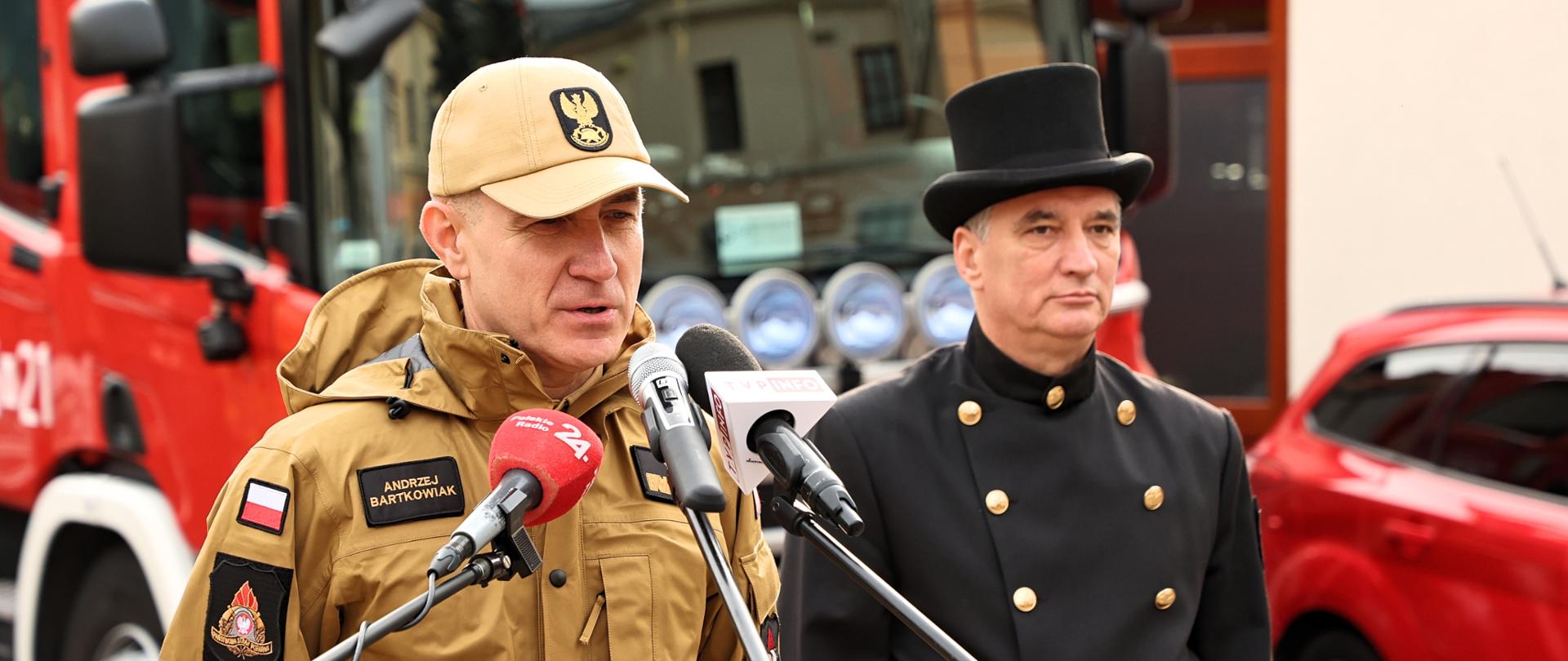Komendant Główny PSP nadbrygadier Andrzej Bartkowiak oraz Pan Mirosław Antosa przedstawiciela Krajowej Izby Kominiarzy podczas briefingu rozpoczynającego ogólnopolską kampanię edukacyjno-informacyjną „Czujka na straży Twojego bezpieczeństwa!” Komendant ubrany w mundur z nakryciem głowy w kolorze piaskowym stoi przed mikrofonami stacji TV i rozgłośni radiowych. Obok komendanta stoi kominiarz w ubraniu galowym. W tle czerwony samochód pożarniczy.