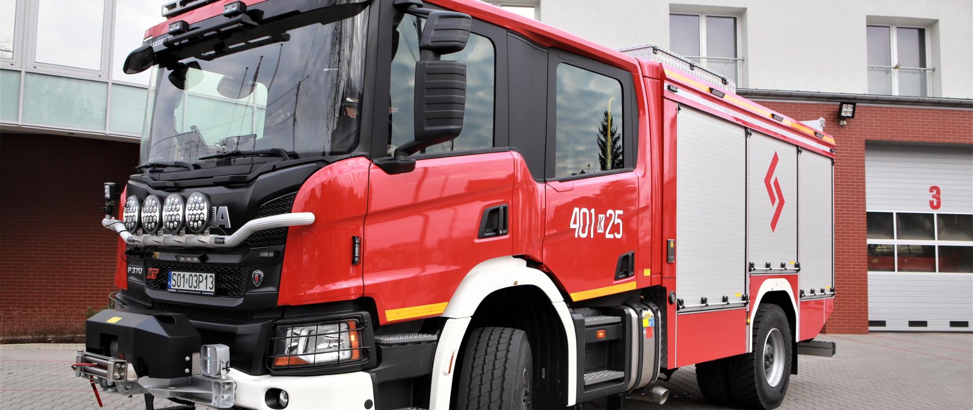 Zdjęcie w centralnej części ukazuje samochód ratowniczo-gaśniczy SCANIA P370. Zdjęcie ukazuje przód i lewą stronę pojazdu. Pojazd stoi na placu zewnętrznym Komendy Powiatowej PSP w Bochni. Samochód jest koloru czerwonego, z czarną maską. Jego skrytki są koloru szarego. Na drugim planie widać budynek komendy powiatowej PSP w Bochni