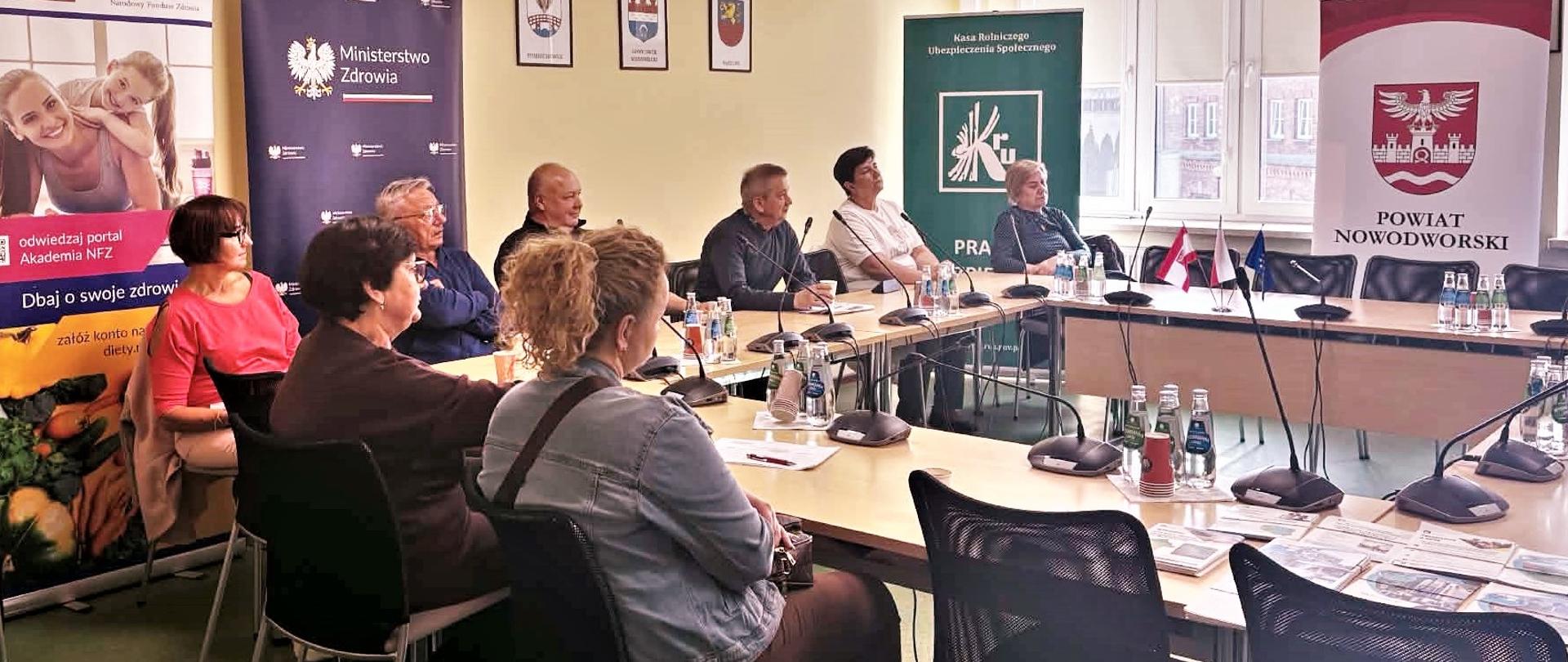 Grupa osób siedzi przy stołach podczas części dotyczącej pytań i analizy zagadnień. Na stołach stoją mikrofony i butelki z wodą, w tle widoczne roll-upy KRUS, NFZ oraz powiatu.