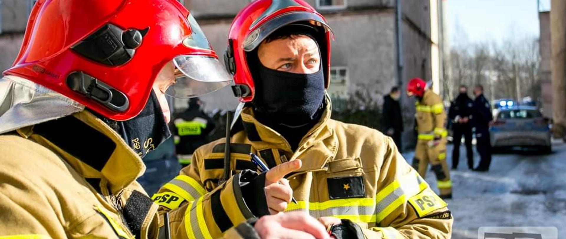 W tle zdjęcia budynki mieszkalne, na pierwszym planie dwóch funkcjonariuszy Państwowej Straży Pożarnej ustalających działania w dłoniach widoczne długopisy. Funkcjonariusze w ubraniach specjalnych koloru piaskowego. W tle widoczni funkcjonariusze policji stojący przed samochodem osobowym oznakowanym - radiowozem. Z uwagi na pandemię osoby na zdjęciu w zabezpieczeniu górnych dróg oddechowym mają maseczki ochronne.